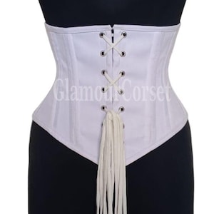 May include: A white corset with a lace-up front and a long, dangling tie. The corset is displayed on a black mannequin. The text "Glamour Corset" is visible on the corset.