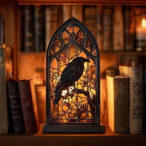 May include: A stained glass lamp featuring a black raven perched on a branch, set against a warm, amber-colored background. The lamp has a gothic arch design and is placed on a wooden surface, surrounded by books.