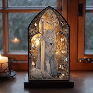 Könnte beinhalten: Dekorative Glasmalerei mit einem Wolf und einem Schwert, umgeben von Schmetterlingen und Blättern. Das Panel ist beleuchtet und erzeugt einen warmen Schein. Das Kunstwerk ist in Schwarz gerahmt und steht auf einem dunklen Holzsockel.