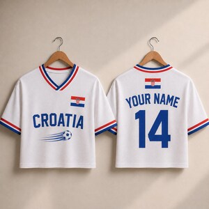 May include: Two white V-neck jerseys with red and blue trim on the sleeves and collar. The front features the text CROATIA, a soccer ball graphic, and the Croatian flag. The back displays the text YOUR NAME and the number 14 with a flag.