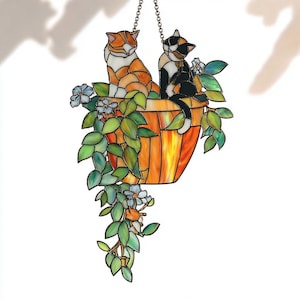May include: Stained glass hanging art featuring two cats in a flower pot. One cat is orange and white, the other is black and white. The pot is orange and yellow with green leaves and blue flowers.