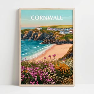 May include: A framed print of Cornwall, featuring a coastal landscape. The artwork depicts a sandy beach, turquoise water, and a village atop a cliff. Wildflowers in shades of pink and orange bloom in the foreground, under a blue sky.