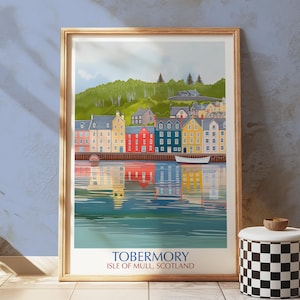 Può includere: Una stampa artistica incorniciata di Tobermory, Isola di Mull, Scozia. L'opera raffigura edifici colorati lungo il lungomare, con riflessi nell'acqua. Una collina verde e un cielo nuvoloso completano la scena. La stampa è in una cornice di legno.