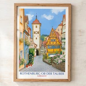 Op de afbeelding: Een ingelijste kunstdruk van een geplaveide straat in Rothenburg ob der Tauber, Duitsland. De scène toont kleurrijke vakwerkhuizen, een hoge klokkentoren en bloeiende bloemen onder een blauwe lucht. De tekst luidt ROTHENBURG OB DER TAUBER GERMANY.