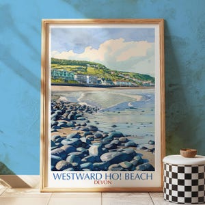 Puede incluir: Una impresión enmarcada de Westward Ho! Beach en Devon, con una ilustración al estilo acuarela. La obra representa una playa con rocas azul-grisáceas, una orilla arenosa y edificios a lo largo de una ladera verde bajo un cielo nublado. El texto "WESTWARD HO! BEACH DEVON" está en la parte inferior.