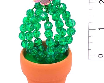 May include: A miniature beaded cactus in a terracotta pot. The cactus is made of green faceted beads and features a pink flower with gold trim. The pot is approximately 2 inches tall.