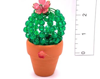 May include: A decorative cactus figurine made of green beads, topped with a pink flower, sits in a small terracotta pot. The cactus is approximately 2 inches tall. The pot has a small pink flower painted on the side.