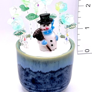 Könnte beinhalten: Eine Keramik-Schneemannfigur mit schwarzem Zylinder, blauem Schal und Besen sitzt in einer blau-weißen Keramikschale. Klare florale Akzente umgeben die Szene, die ein Winterthema darstellt.