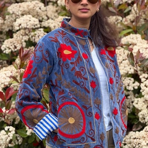 Peut inclure: Veste en velours bleu avec broderies florales rouges et marron. La veste présente un col montant, une fermeture éclair et des poignets rayés. Le design comprend des motifs circulaires et floraux, créant un look orné.