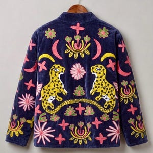 May include: Navy blue velvet jacket with colourful embroidered details. The jacket features two yellow leopard figures, floral and geometric patterns in pink, yellow, and green. The jacket is hanging on a wooden hanger.