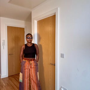 Puede incluir: Una persona con una camiseta sin mangas negra y pantalones de pernera ancha con rayas verticales en tonos naranja, morado y blanco. Los pantalones tienen bolsillos de parche y cintura con cordón. La persona lleva gafas de sol negras y zapatos negros.