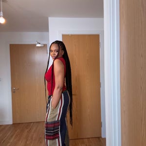 May include: A person wearing a red sleeveless top and striped pants with a pocket. The pants have vertical stripes in beige, navy, and red. The person has long black braids.