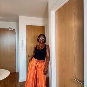 Puede incluir: Una camiseta sin mangas negra combinada con pantalones anchos a rayas naranjas. Los pantalones tienen un cinturón estampado. La persona lleva gafas de sol, un collar y una pulsera dorados. El atuendo es informal y elegante.