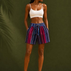 May include: A woman wearing a white crop top and striped shorts. The shorts have vertical stripes in blue, pink, and purple. The shorts have a drawstring waist. The background is a solid green.