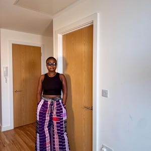 Puede incluir: Una persona con un top corto negro y pantalones de pernera ancha. Los pantalones presentan un vibrante estampado geométrico morado, rojo y blanco. El atuendo se combina con zapatos negros y gafas. El fondo incluye puertas de madera y paredes blancas.