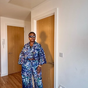 Könnte beinhalten: Ein zweiteiliges Set in Blau-, Grün- und Weißtönen, bestehend aus einer cropped Jacke und einer Hose mit weitem Bein. Das Set hat ein von einer Stadt inspiriertes Muster. Die Jacke hat weite Ärmel und die Hose eine hohe Taille. Das Outfit wird mit Sonnenbrille getragen.