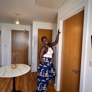 Puede incluir: Una persona lleva un top blanco con cuello halter y pantalones de pernera ancha de cintura alta con un estampado geométrico azul y blanco. Los pantalones tienen bolsillos. La persona sonríe y se apoya en una puerta de madera en una habitación bien iluminada.
