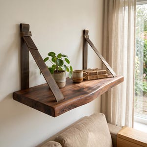 Op de afbeelding: Een zwevend houten plank met een natuurlijke rand, opgehangen aan bruine leren riemen. Het toont een kleine potplant, een keramische mok en boeken, wat een minimalistische en natuurlijke sfeer creëert.