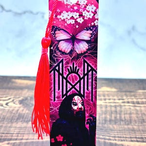 May include: A pink bookmark with a butterfly and floral design at the top, transitioning to a graphic image of a figure in a black cloak. A red tassel hangs from the top. The bookmark is a unique gift.