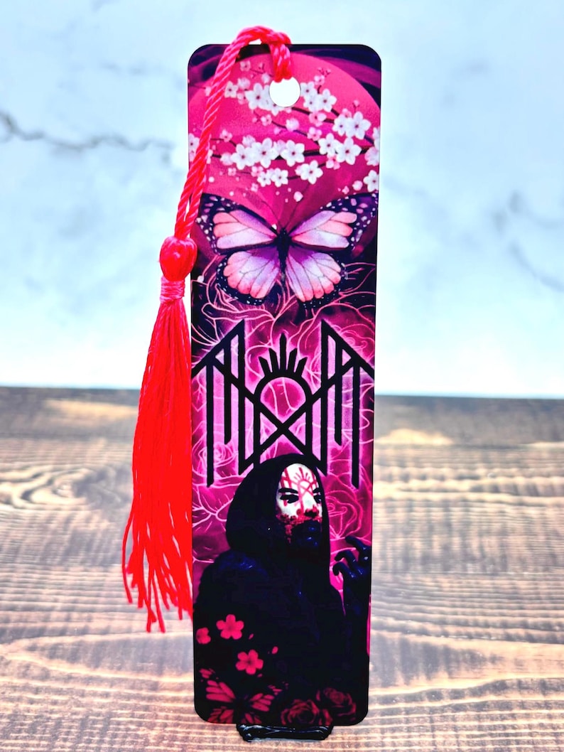 May include: A pink bookmark with a butterfly and floral design at the top, transitioning to a graphic image of a figure in a black cloak. A red tassel hangs from the top. The bookmark is a unique gift.