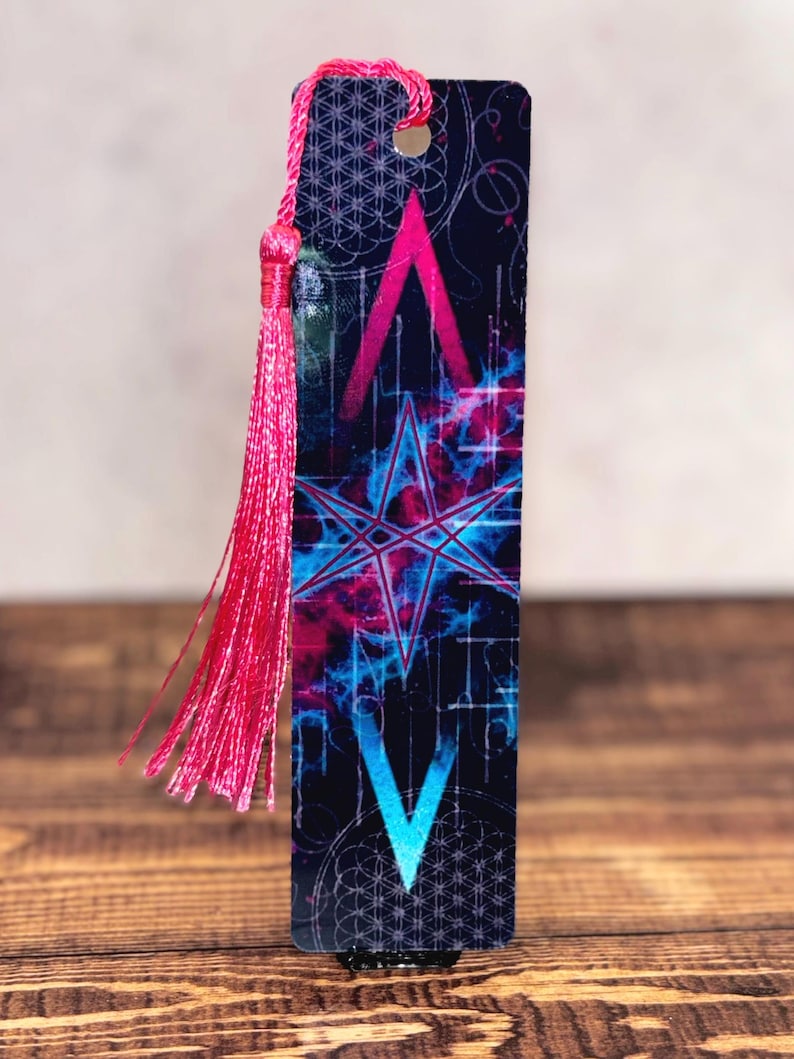May include: A rectangular bookmark with a dark blue background featuring a pink and blue star design. The bookmark has a pink tassel attached to the top. The design includes geometric shapes and a celestial theme.