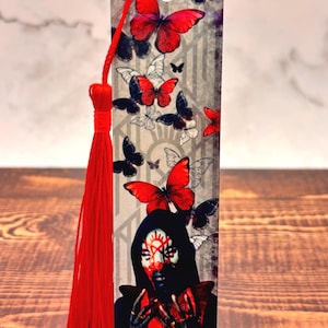 May include: A decorative bookmark featuring a design of red and black butterflies, with a central image of a person with face paint. The bookmark has a red tassel attached at the top.
