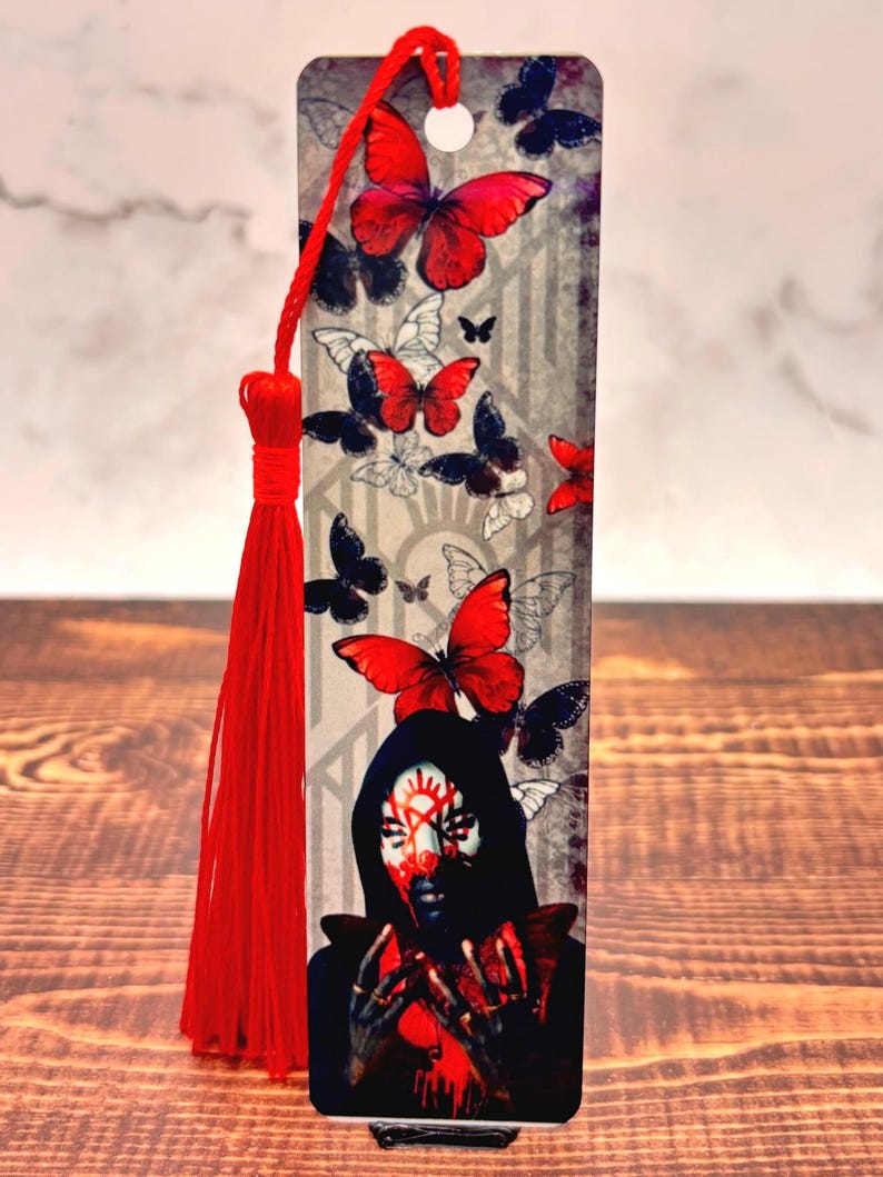 May include: A decorative bookmark featuring a design of red and black butterflies, with a central image of a person with face paint. The bookmark has a red tassel attached at the top.