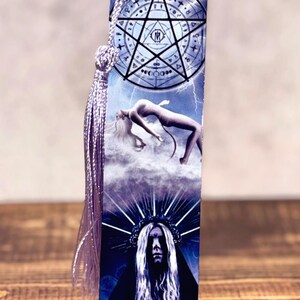 May include: A rectangular bookmark with a purple tassel and cord. The bookmark features a celestial design with a pentagram, a figure floating in clouds, and a portrait of a person with a crown. The background is blue with lightning.