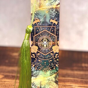 May include: A rectangular bookmark with a forest scene design, featuring a central geometric pattern with gold accents and floral details. A green tassel hangs from the top. The bookmark has a hole at the top for the tassel.