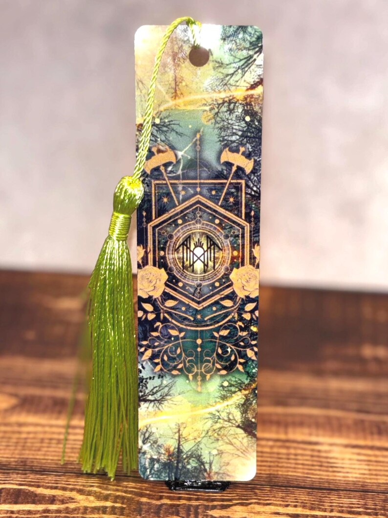 May include: A rectangular bookmark with a forest scene design, featuring a central geometric pattern with gold accents and floral details. A green tassel hangs from the top. The bookmark has a hole at the top for the tassel.