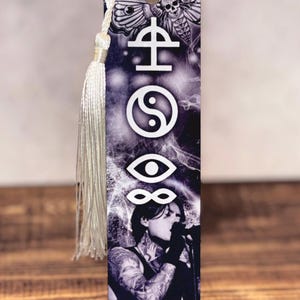 May include: A black and white bookmark featuring a singer with tattoos, symbols, and a moth with a skull design. The bookmark has a silver tassel attached to the top with a white cord.