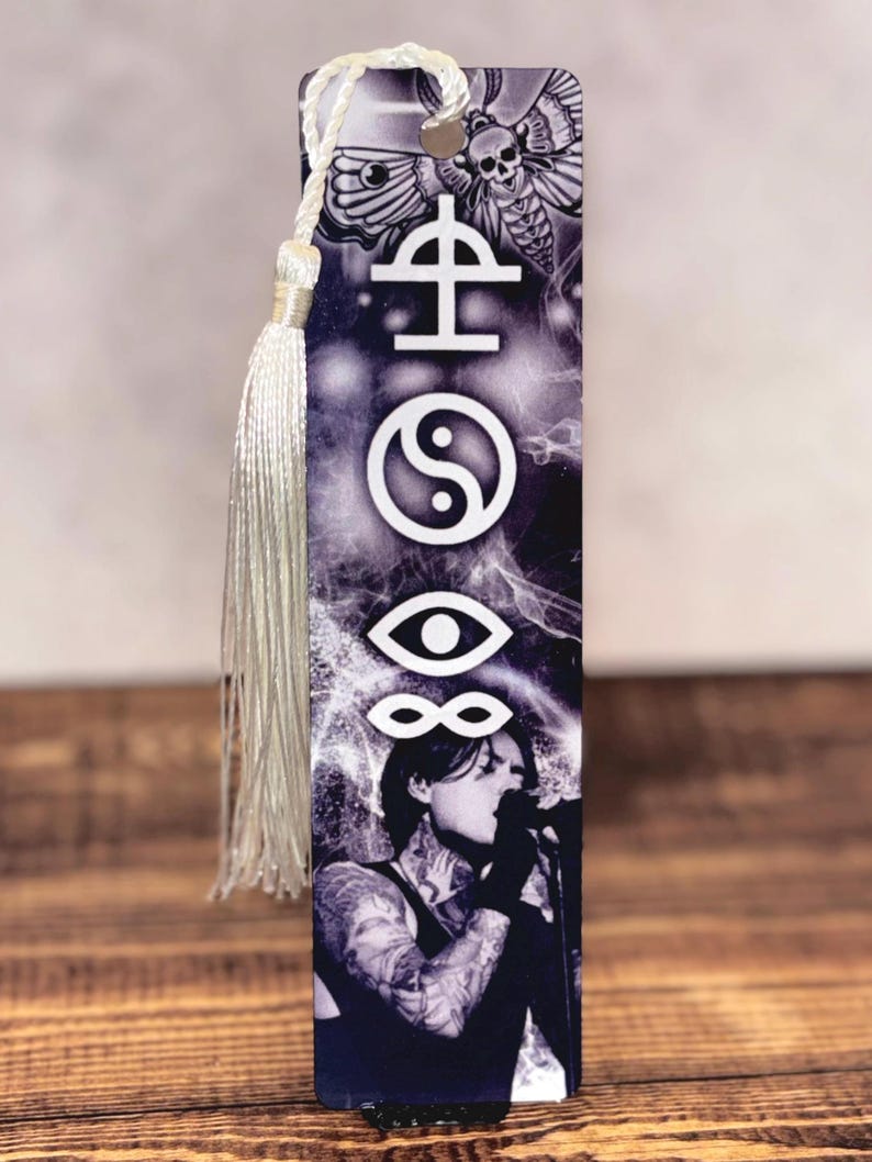 May include: A black and white bookmark featuring a singer with tattoos, symbols, and a moth with a skull design. The bookmark has a silver tassel attached to the top with a white cord.