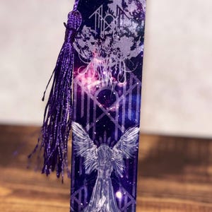 May include: A rectangular bookmark with a celestial design. The bookmark features a purple tassel and a detailed illustration of an angel with outstretched wings. The background is a deep blue with a starry night sky and geometric patterns.