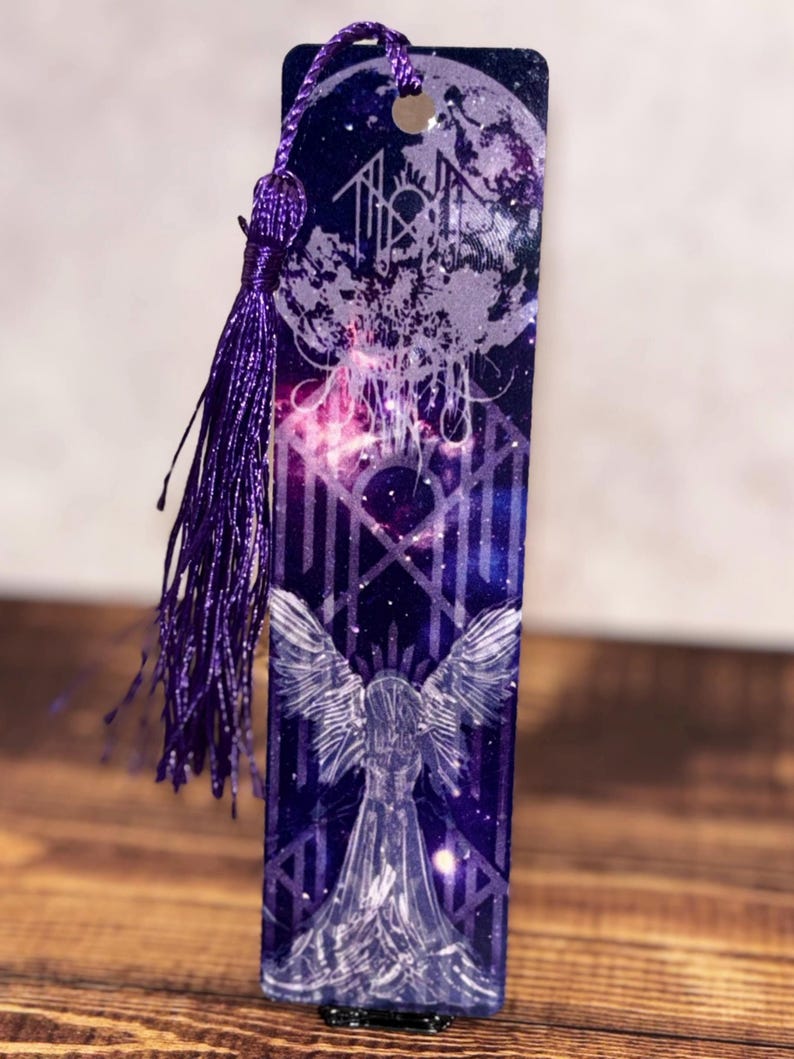 May include: A rectangular bookmark with a celestial design. The bookmark features a purple tassel and a detailed illustration of an angel with outstretched wings. The background is a deep blue with a starry night sky and geometric patterns.