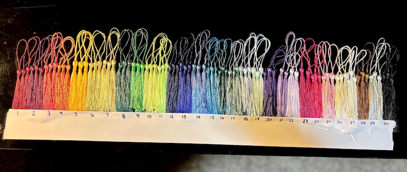 May include: A collection of 30 colorful tassels arranged in a rainbow spectrum, each with a looped string for hanging. The tassels range in color from red to black, with a white numbered guide below each tassel.