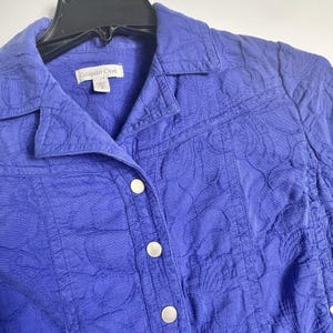 May include: A close-up of a periwinkle blue jacket with a floral pattern. The jacket has a collared neckline, three white buttons, and the brand name "Coldwater Creek" on the label.