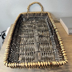 May include: Rectangular woven wicker basket with a handle. The basket is mainly dark grey with lighter brown accents on the rim and handle. The basket is empty and sits on a light brown surface.