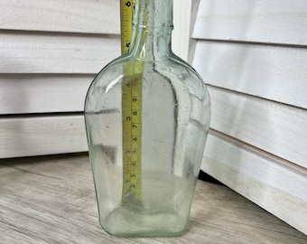 May include: A clear, antique-style glass bottle with a rounded body and a narrow neck. A yellow measuring tape is placed inside the bottle, showing a height of approximately 7 inches. The bottle has a slight green tint and is set against a white shutter background.