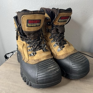 Puede incluir: Par de botas de trabajo aisladas Rocky Thinsulate Ultra. Las botas son de cuero marrón y beige con suelas de goma negra. Las botas tienen cordones negros y marrones y ojales metálicos. El logotipo de Rocky es visible.