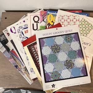 Puede incluir: Una colección de libros de patrones de acolchado, con el patrón "Feelin' Groovy Quilt" en primer plano, que muestra un diseño de acolchado geométrico con hexágonos azules, morados y estampados. Otros títulos incluyen "Quilt" y "Needlework".