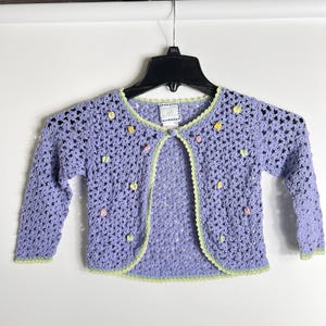 May include: A lavender crochet cardigan with long sleeves, accented with small, colorful flower-shaped embellishments. The edges are trimmed with a light green scalloped design. The open-front sweater is hanging on a black hanger.
