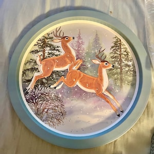 May include: Round serving tray with a light blue rim. The tray features a winter scene with two leaping deer in a snowy forest. The deer are brown with white spots and antlers. The background includes evergreen trees and falling snow.