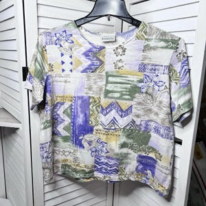 May include: A short-sleeved shirt with a patchwork design in shades of lavender, green, and beige. The shirt features floral and geometric patterns. The tag reads "Editions".