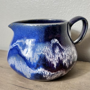 May include: A ceramic pitcher with a dark blue handle and spout. The body of the pitcher features a blend of blue, white, and dark purple hues, creating a marbled effect. The pitcher is sitting on a wooden surface.