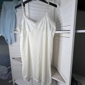 以下が含まれることがあります： アイボリー色のスリップドレス。ネックラインと裾に繊細なレースのトリムが施されています。ドレスは調節可能なスパゲッティストラップとVネックのデザインが特徴です。スリップは、白い折りたたみ式のスクリーンを背景に、黒いハンガーに吊るされています。