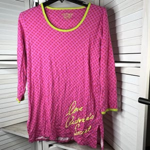 Può includere: Top rosa a maniche lunghe con motivo a stelle e finiture verde neon. La parte superiore presenta il testo "Victoria's Secret" sul décolleté e "Love Victoria's Secret" in corsivo sul davanti. La camicia ha un orlo spaccato.