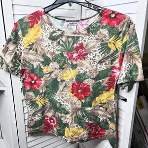 May include: A short-sleeved t-shirt with a vibrant floral print. The shirt features a pattern of red hibiscus flowers, yellow flowers, and green leaves on a tan background. The word "EDITIONS" is visible on the neckline.