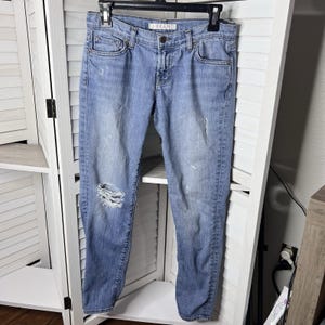 Puede incluir: Vaqueros de mezclilla azul claro con detalles desgastados, incluyendo un gran rasgón en la rodilla izquierda. Los vaqueros están colgados de una percha negra. La marca "J BRAND" es visible en la cintura.