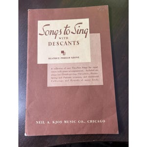 May include: A vintage songbook titled "Songs to Sing with Descants" by Beatrice Perham Krone. The cover is a muted brown with a white rectangular inset containing the title and author. The book includes songs for various occasions.