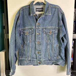 May include: A medium-wash blue denim jacket featuring a classic collar, long sleeves, and a front button closure. The garment includes two chest pockets with buttoned flaps and two side slit pockets. A label reading LA LOVING is at the neck.